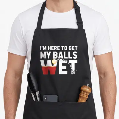 Wet beer pong apron - White stacked text reading I'M HERE TO GET MY BALLS WET with red cups and yellow ball trajectory — shown on a black cotton canvas apron against a white background