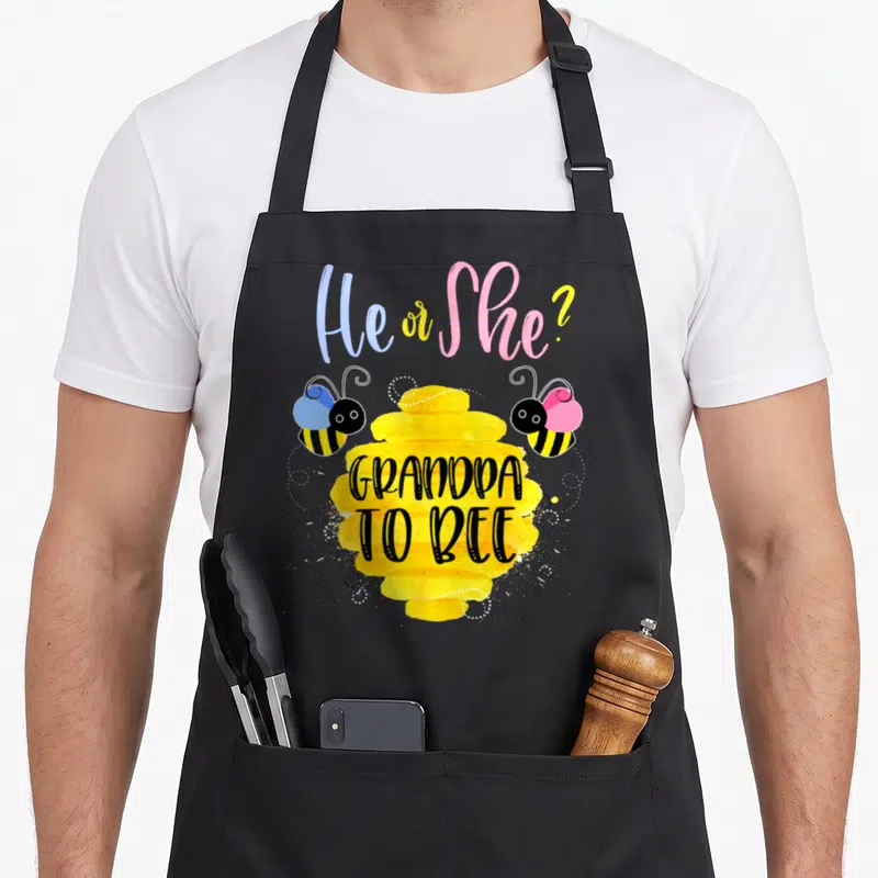 Grandpa bee apron - He or She text over yellow honeycomb flanked by blue and pink cartoon bees — shown on a black cotton canvas apron against a white background