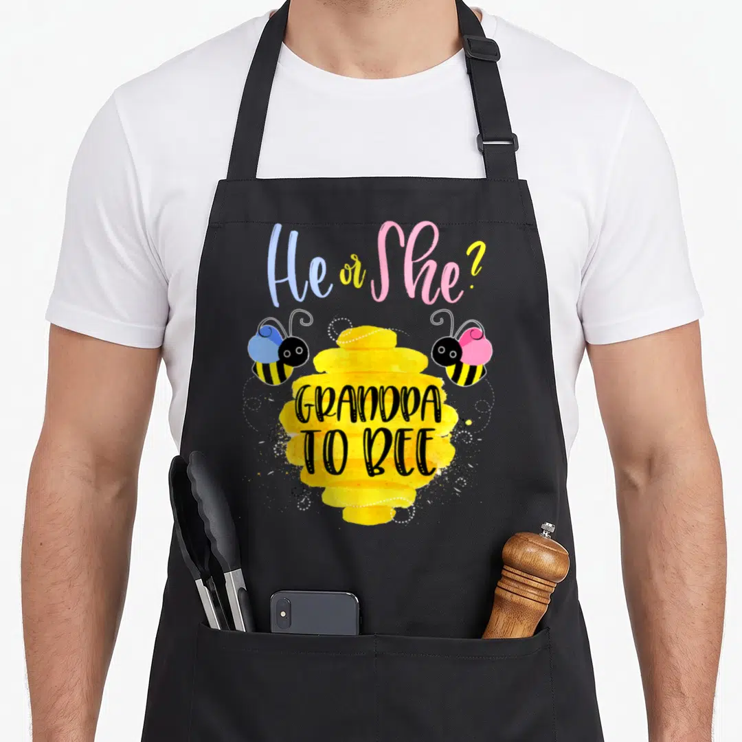 Grandpa bee apron - He or She text over yellow honeycomb flanked by blue and pink cartoon bees — shown on a black cotton canvas apron against a white background