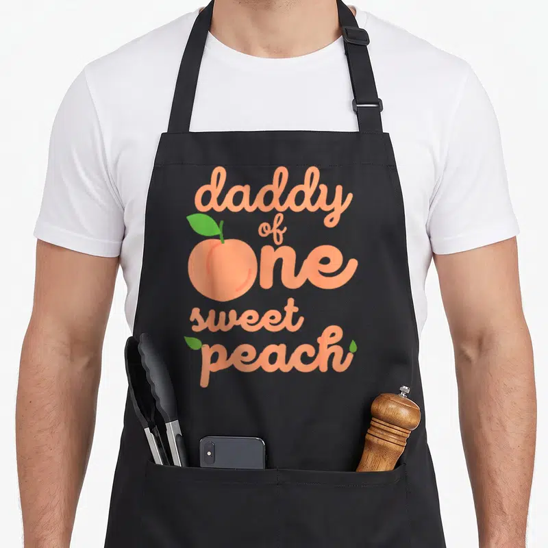 Peach daddy apron - daddy of one sweet peach text with peach fruit replacing the letter O — shown on a black cotton canvas apron against a white background