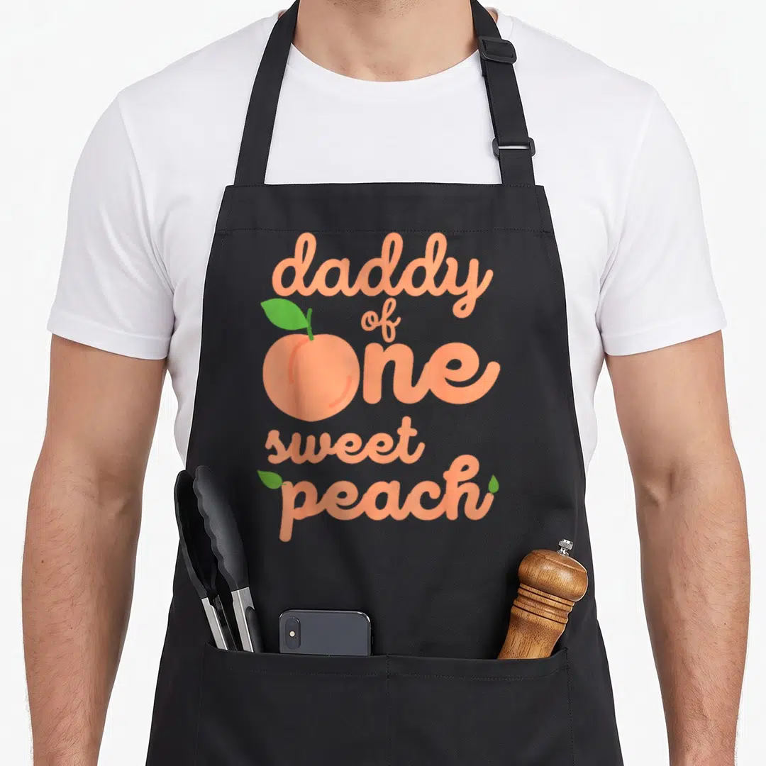 Peach daddy apron - daddy of one sweet peach text with peach fruit replacing the letter O — shown on a black cotton canvas apron against a white background