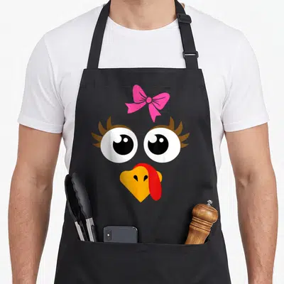 Vintage turkey thanksgiving day aprons - Cartoon turkey face with pink bow, big eyes, and orange beak on black — shown on a black cotton canvas apron against a white background
