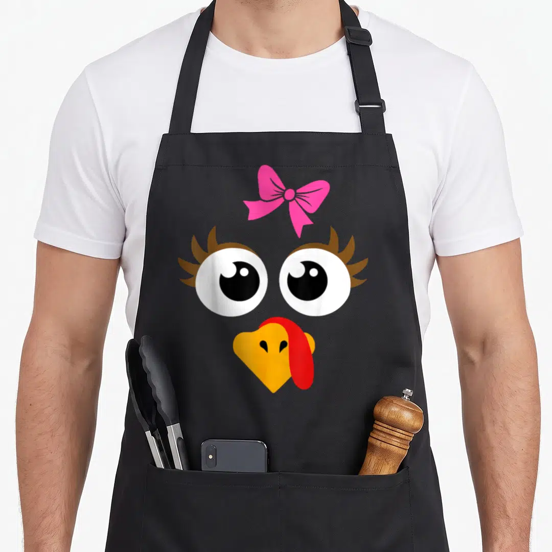 Vintage turkey thanksgiving day aprons - Cartoon turkey face with pink bow, big eyes, and orange beak on black — shown on a black cotton canvas apron against a white background