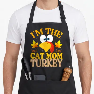 Cat funny turkey apron - Cartoon turkey head between I'M THE CAT MOM TURKEY text and two autumn leaves — shown on a black cotton canvas apron against a white background