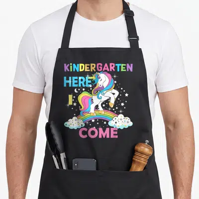 Come unicorn aprons - White unicorn on rainbow with stars and text KINDERGARTEN HERE I COME — shown on a black cotton canvas apron against a white background