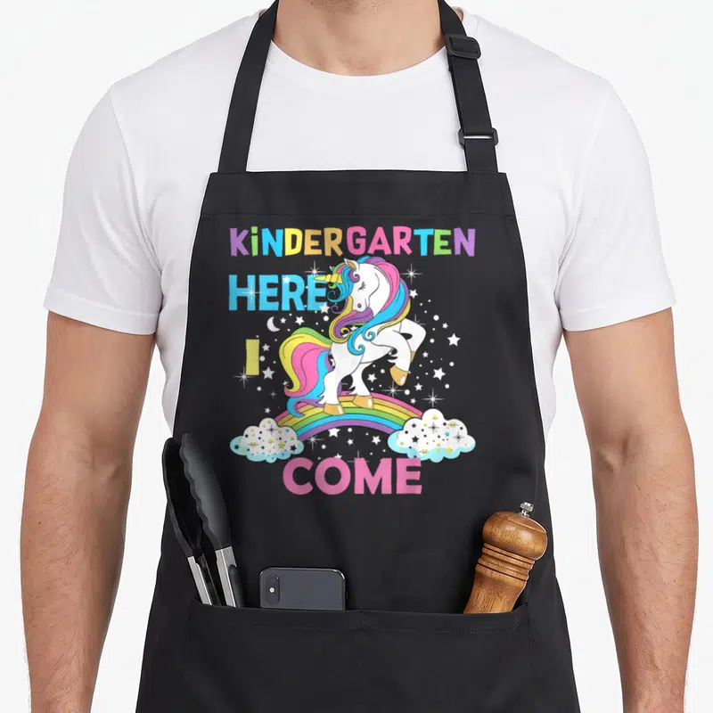 Come unicorn aprons - White unicorn on rainbow with stars and text KINDERGARTEN HERE I COME — shown on a black cotton canvas apron against a white background
