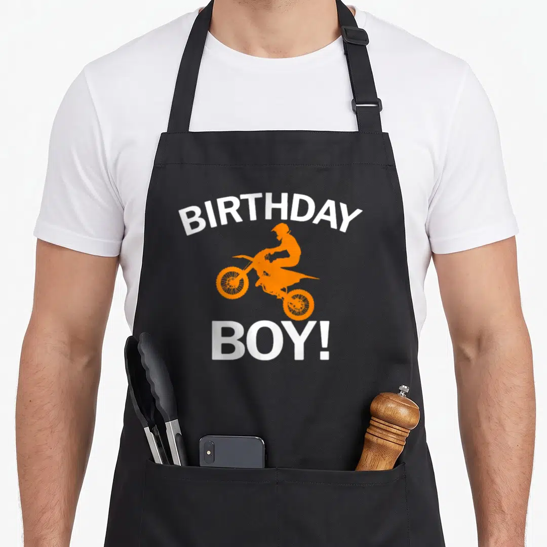 Dirt birthday apron - White BIRTHDAY BOY text with orange dirt bike rider silhouette on grey background — shown on a black cotton canvas apron against a white background