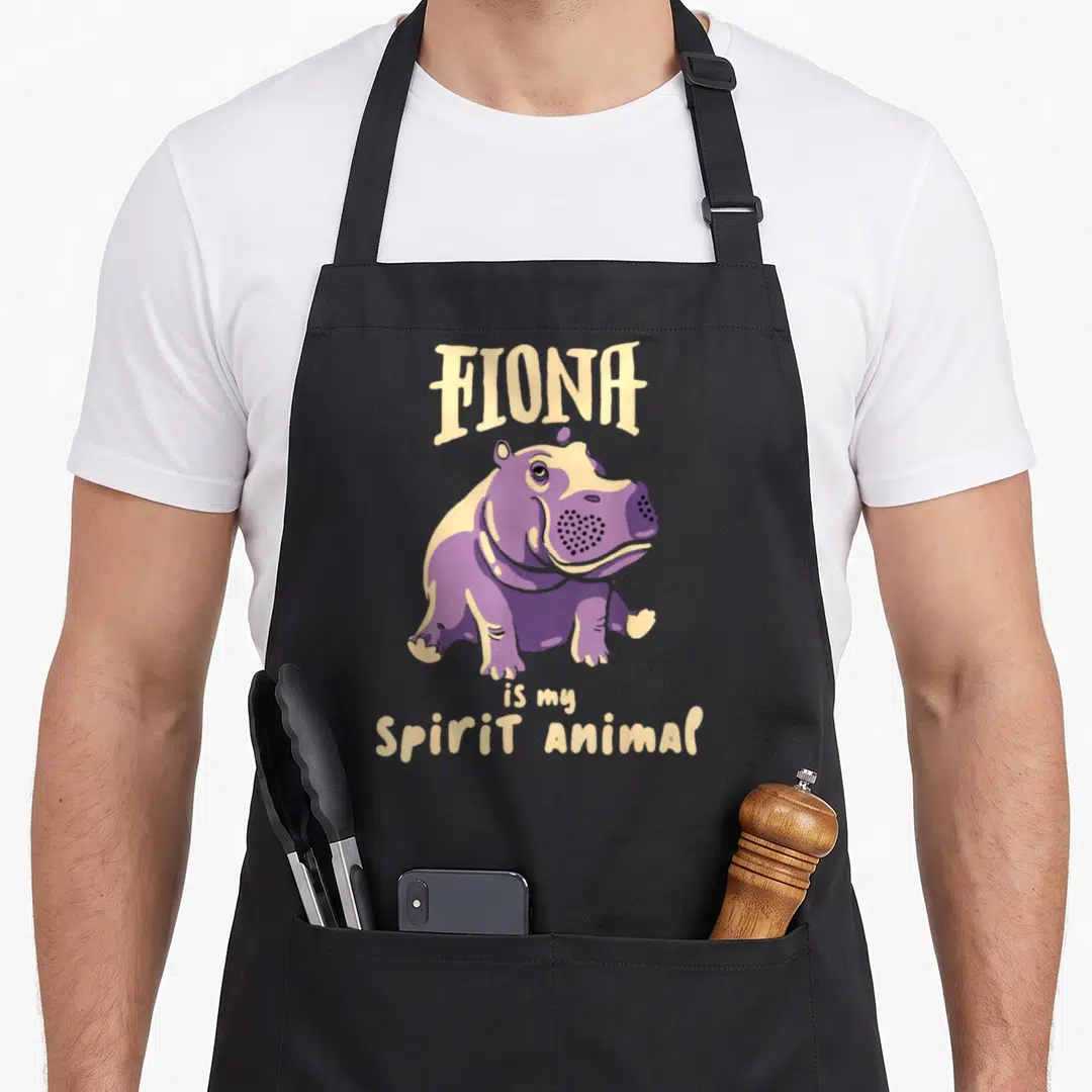 Fiona animal apron - Purple baby hippo illustration with stacked text FIONA is my Spirit Animal — shown on a black cotton canvas apron against a white background
