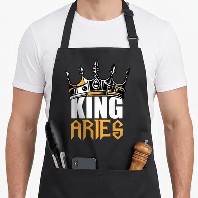 King aries kitchen apron - Black bib apron with crown graphic and KING ARIES text in white and orange — shown on a black cotton canvas apron against a white background