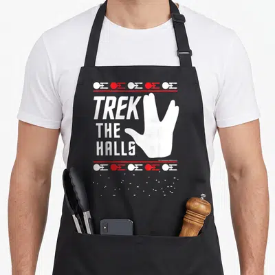 Hall christmas sweater apron - White Vulcan salute hand beside stacked text TREK THE HALLS framed by red lines — shown on a black cotton canvas apron against a white background