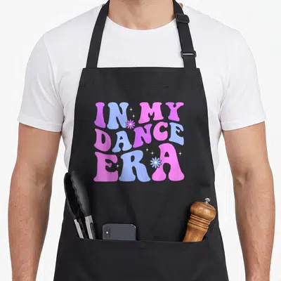 Era ballet dance apron - Pink purple and blue IN MY DANCE ERA typography with small flowers and stars — shown on a black cotton canvas apron against a white background