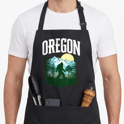 Oregon bigfoot tree apron - Retro OREGON graphic with Bigfoot silhouette, mountains, trees, and yellow sun in green — shown on a black cotton canvas apron against a white background