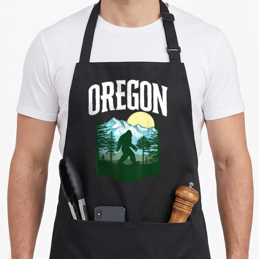 Oregon bigfoot tree apron - Retro OREGON graphic with Bigfoot silhouette, mountains, trees, and yellow sun in green — shown on a black cotton canvas apron against a white background