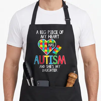 Awareness mom daughter apron - Distressed heart with multicolor puzzle pieces and text about autism and daughter — shown on a black cotton canvas apron against a white background