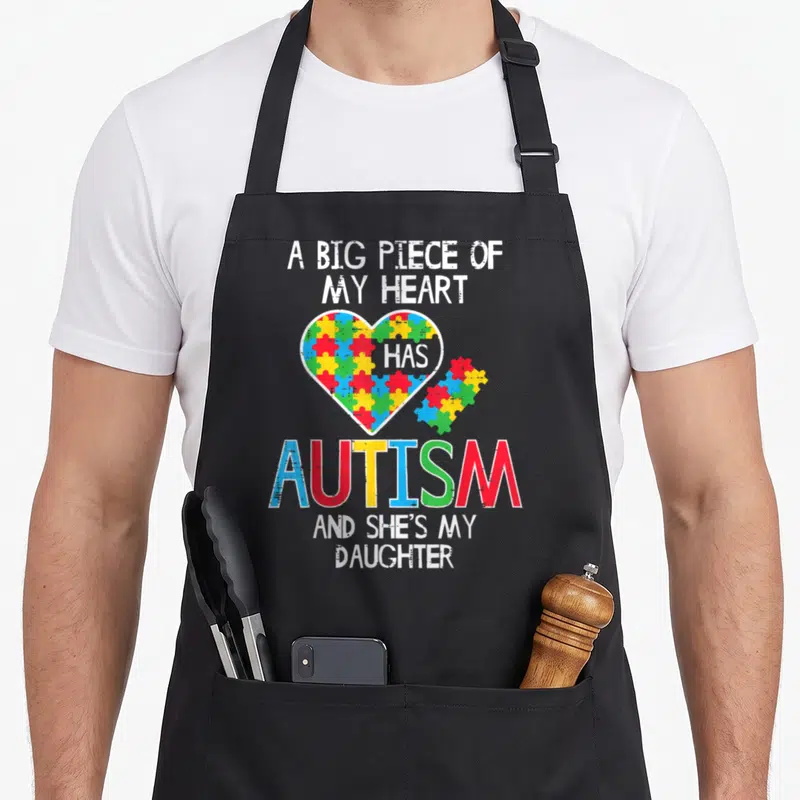Awareness mom daughter apron - Distressed heart with multicolor puzzle pieces and text about autism and daughter — shown on a black cotton canvas apron against a white background