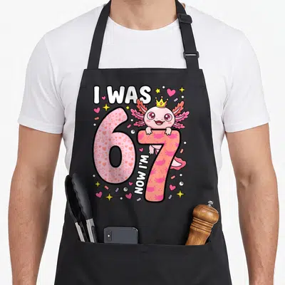 Axolotl 7th birthday apron - Pink axolotl with crown peeking over number 7 with I WAS 6 NOW I'M 7 text and confetti — shown on a black cotton canvas apron against a white background