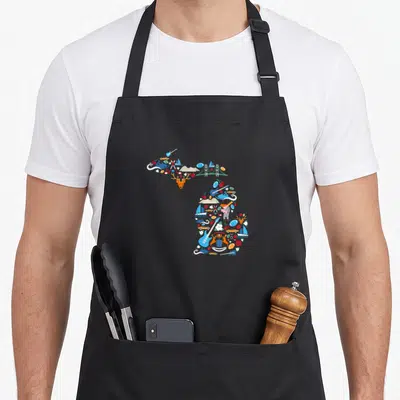 Icon pride apron - Map of Michigan composed of cartoon icons including guitars, deer, sailboats, and cherries in blue — shown on a black cotton canvas apron against a white background