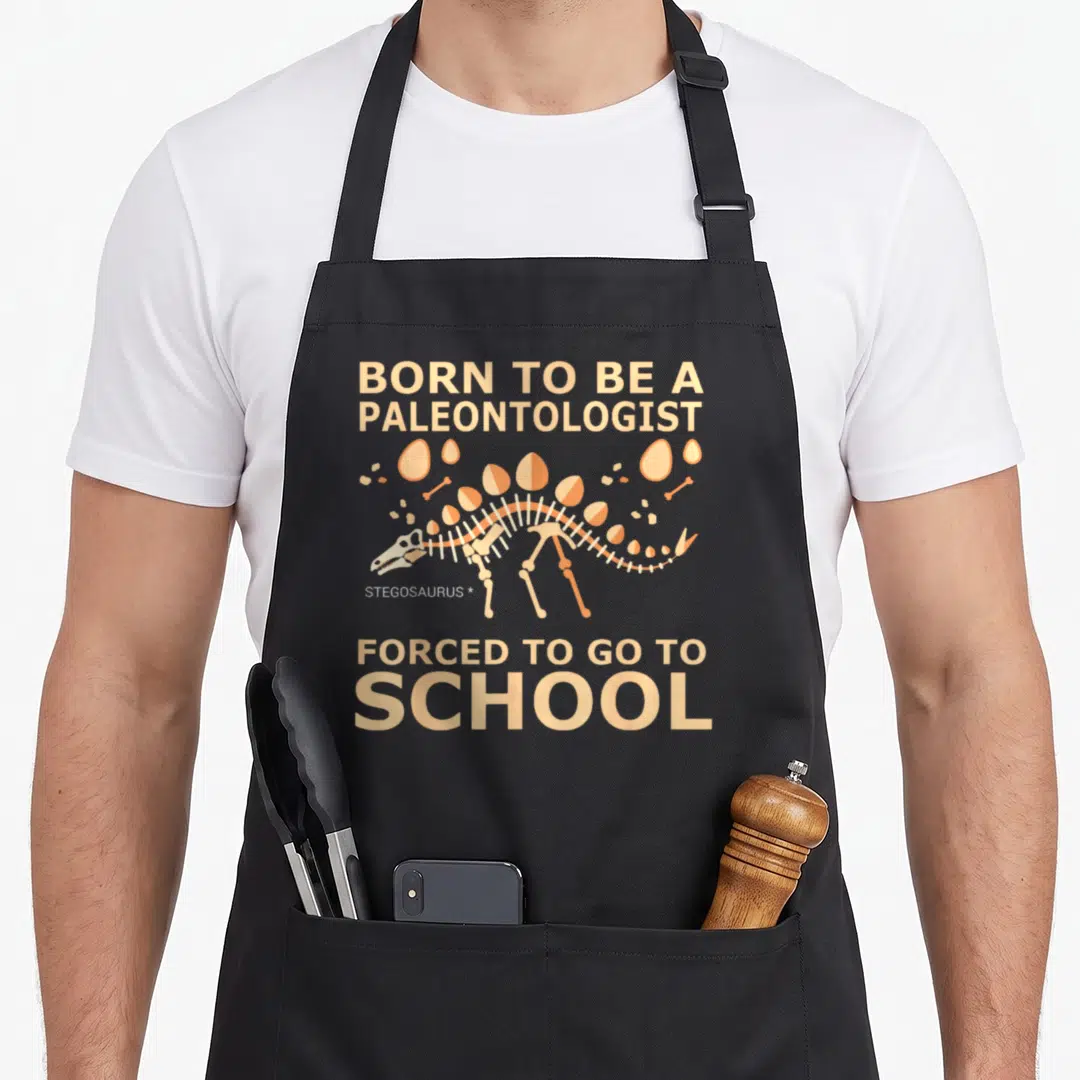 Paleontologist forced school apron - Stegosaurus skeleton with eggs and bones in peach and white under printed text — shown on a black cotton canvas apron against a white background