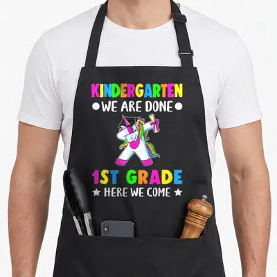 Kindergarten grade come and get it apron - Dabbing unicorn with rainbow text reading we are done 1st grade here we come — shown on a black cotton canvas apron against a white background