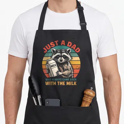 Alway came raccoon milk aprons - Raccoon holding milk carton in front of red yellow and teal striped sunset circle — shown on a black cotton canvas apron against a white background