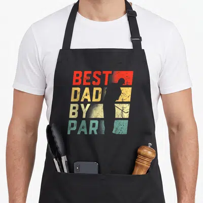 Par golf father day apron - Stacked BEST DAD BY PART text with golfer silhouette in red yellow teal and grey — shown on a black cotton canvas apron against a white background