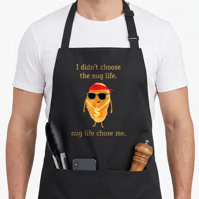 Nugget chicken print apron - Cartoon nugget wearing red bandana, sunglasses, and gold dollar chain with punny text — shown on a black cotton canvas apron against a white background