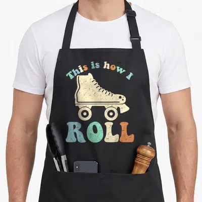 Roll 70s apron - Retro cream roller skate with arched multicolor text reading This is how I ROLL — shown on a black cotton canvas apron against a white background