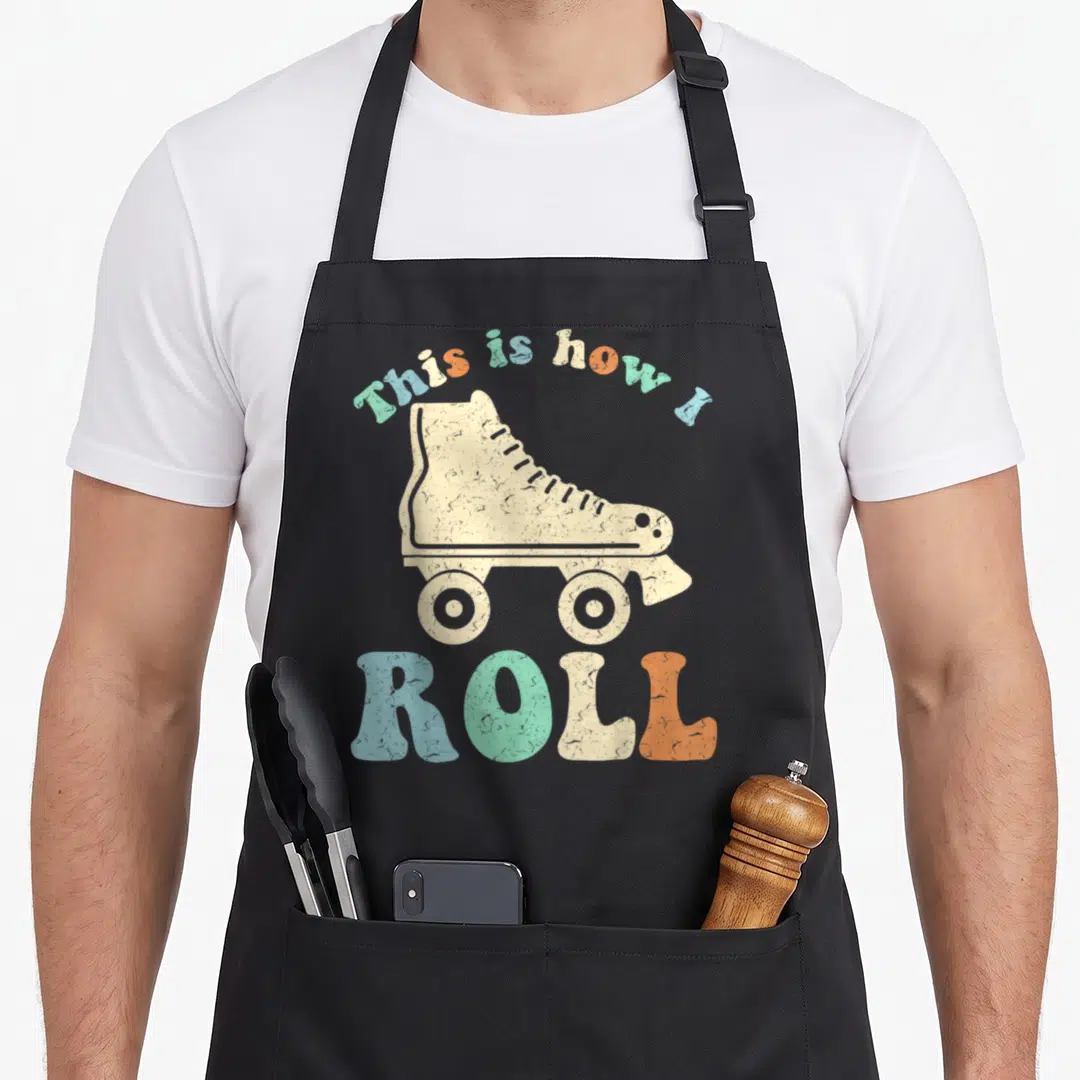 Roll 70s apron - Retro cream roller skate with arched multicolor text reading This is how I ROLL — shown on a black cotton canvas apron against a white background