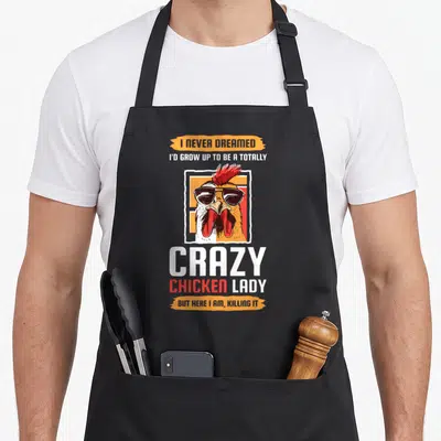 Totally chicken apron - Cartoon rooster in sunglasses on orange and white background with humorous text about being — shown on a black cotton canvas apron against a white background