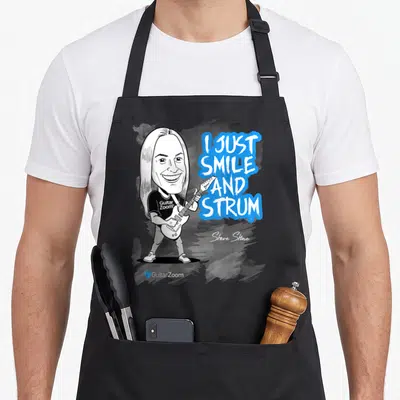 Steve caricature apron - Cartoon guitarist next to I Just Smile And Strum text on blue splash with Steve Stine signature — shown on a black cotton canvas apron against a white background