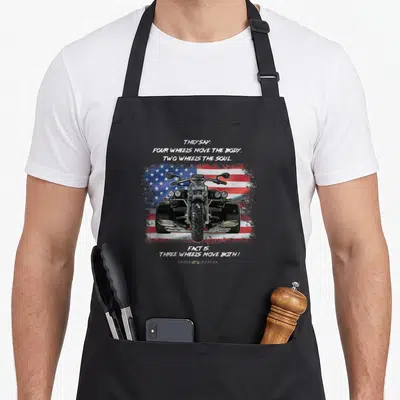 Three wheel apron - Realistic trike on distressed US flag with quote about three wheels moving both body and soul — shown on a black cotton canvas apron against a white background