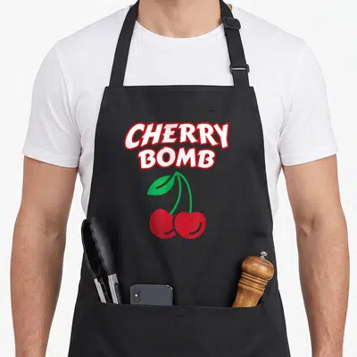 70s bomb cherry apron - Stacked CHERRY BOMB text above two red cherries with green leaf — shown on a black cotton canvas apron against a white background