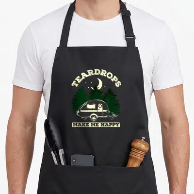 Teardrop trailer apron - Vintage camper illustration with pine trees and stacked Teardrops Make Me Happy text — shown on a black cotton canvas apron against a white background