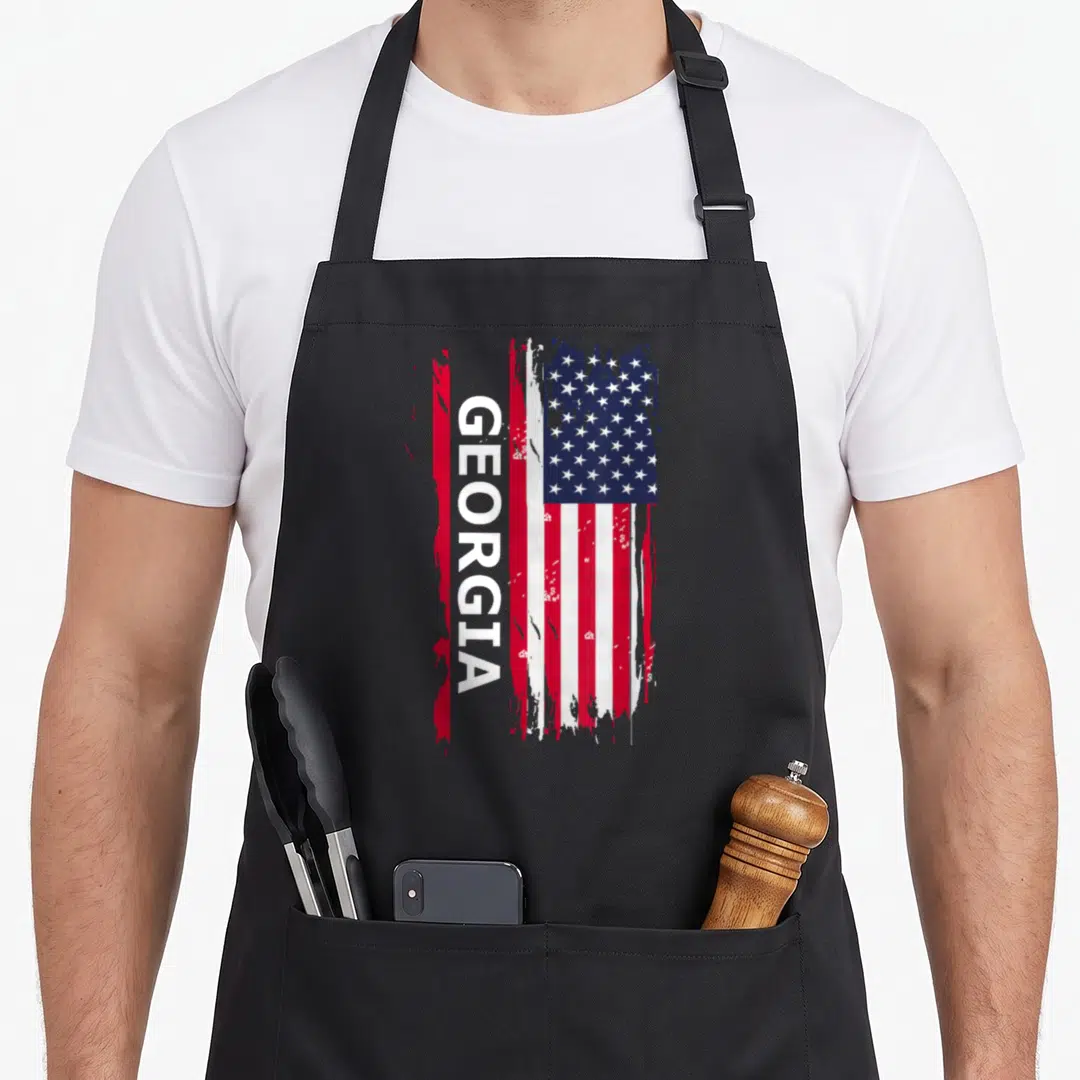 Funny georgia apron - Vertical GEORGIA text over distressed red white blue flag — shown on a black cotton canvas apron against a white background