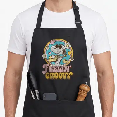 Feelin groovy apron - Snoopy playing guitar inside blue circle with hearts and flowers, stacked FEELIN GROOVY text below — shown on a black cotton canvas apron against a white background
