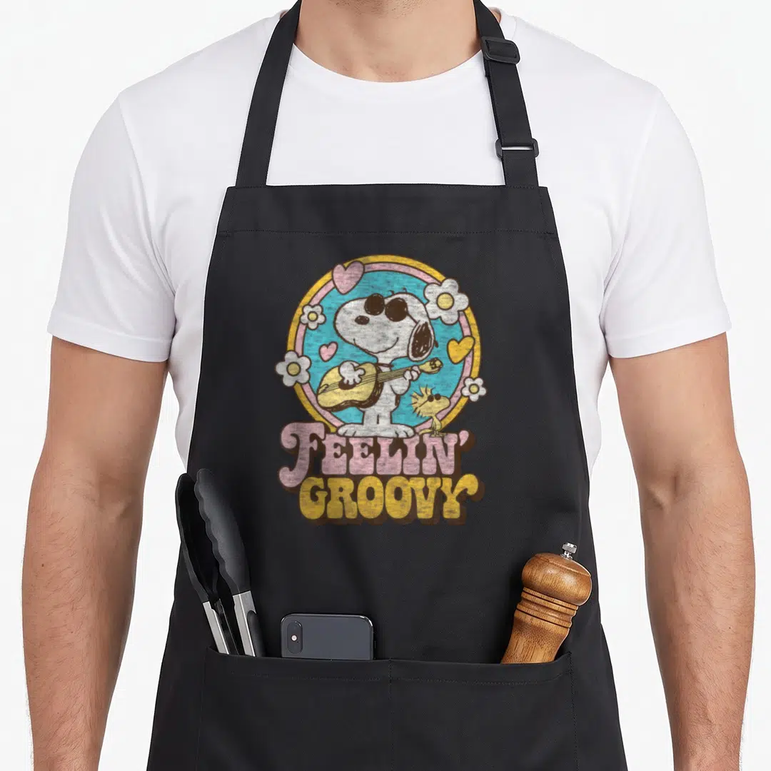 Feelin groovy apron - Snoopy playing guitar inside blue circle with hearts and flowers, stacked FEELIN GROOVY text below — shown on a black cotton canvas apron against a white background