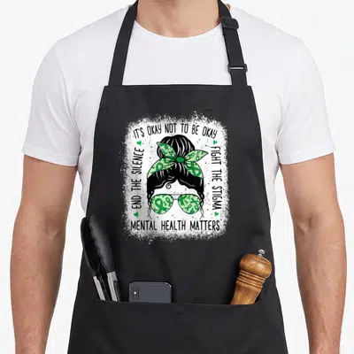 Mental messy aprons - Cartoon woman with teal ribbon sunglasses and IT'S OKAY NOT TO BE OKAY text — shown on a black cotton canvas apron against a white background