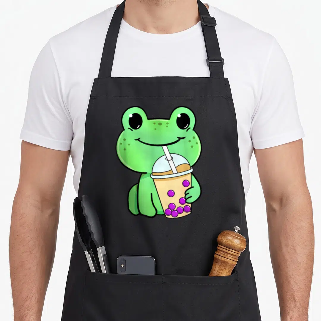 Drinking tea apron - Cartoon green frog holding a bubble tea cup with purple pearls and a straw — shown on a black cotton canvas apron against a white background