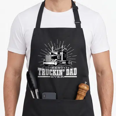 Truckin trucking truck apron - White semi-truck illustration above stacked best truckin dad ever text on black — shown on a black cotton canvas apron against a white background