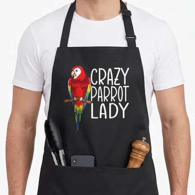 Talking ornithology bird apron - Cartoon red parrot perched on branch beside vertical white text reading crazy parrot — shown on a black cotton canvas apron against a white background