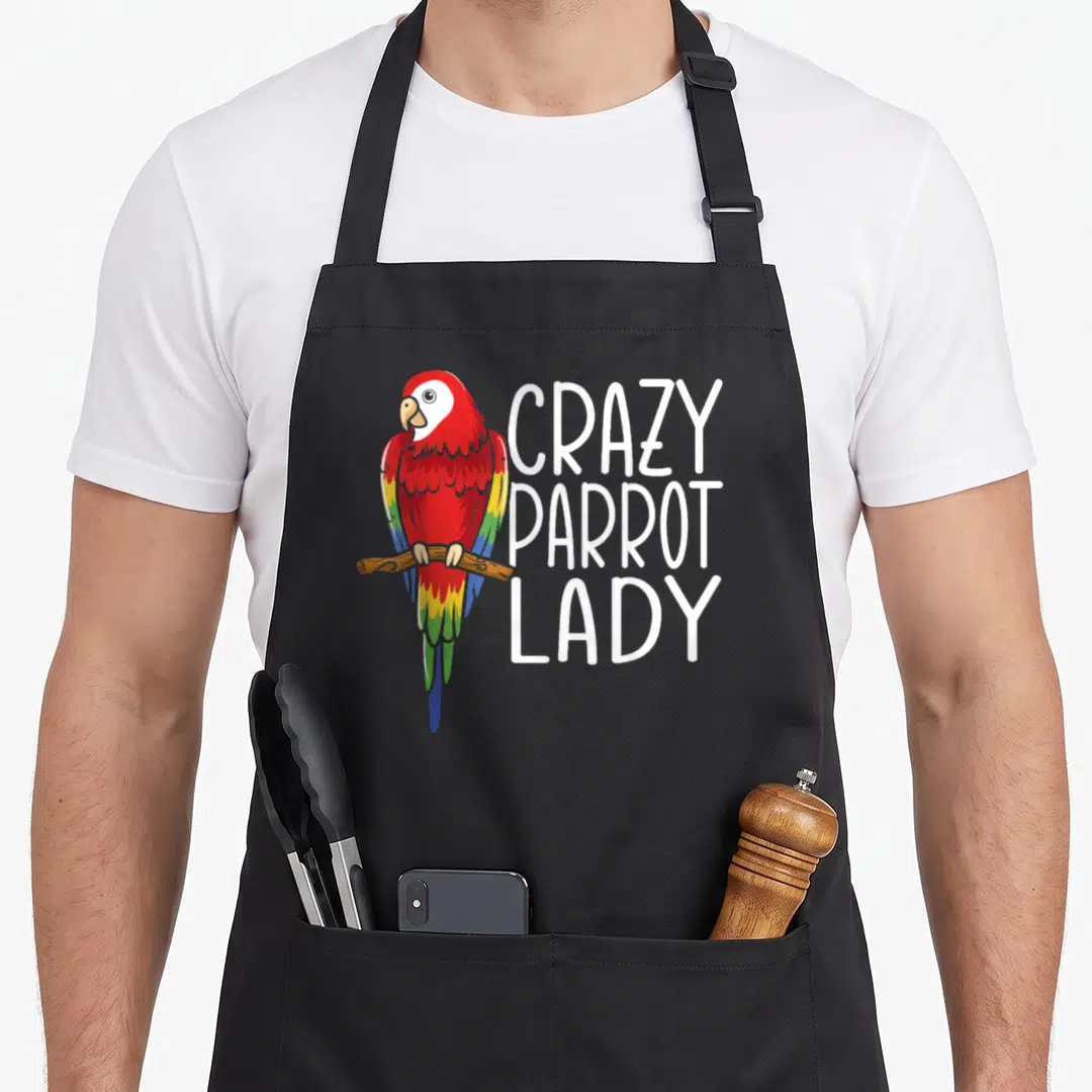 Talking ornithology bird apron - Cartoon red parrot perched on branch beside vertical white text reading crazy parrot — shown on a black cotton canvas apron against a white background