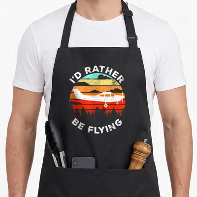Airplane flying apron - Retro sunset with white airplane, pine trees, and text I'D RATHER BE FLYING — shown on a black cotton canvas apron against a white background