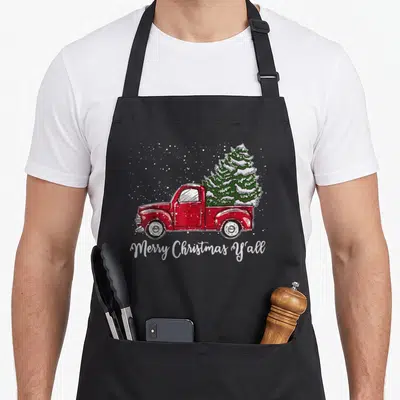 Truck merry christmas apron - Red truck carrying snow-covered green tree with white Merry Christmas Y'all text — shown on a black cotton canvas apron against a white background