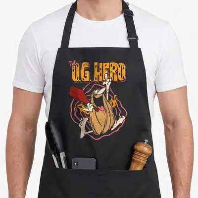 Caveman captain apron - Cartoon caveman holding red club under arched orange text THE U.G. HERO on pink concentric — shown on a black cotton canvas apron against a white background