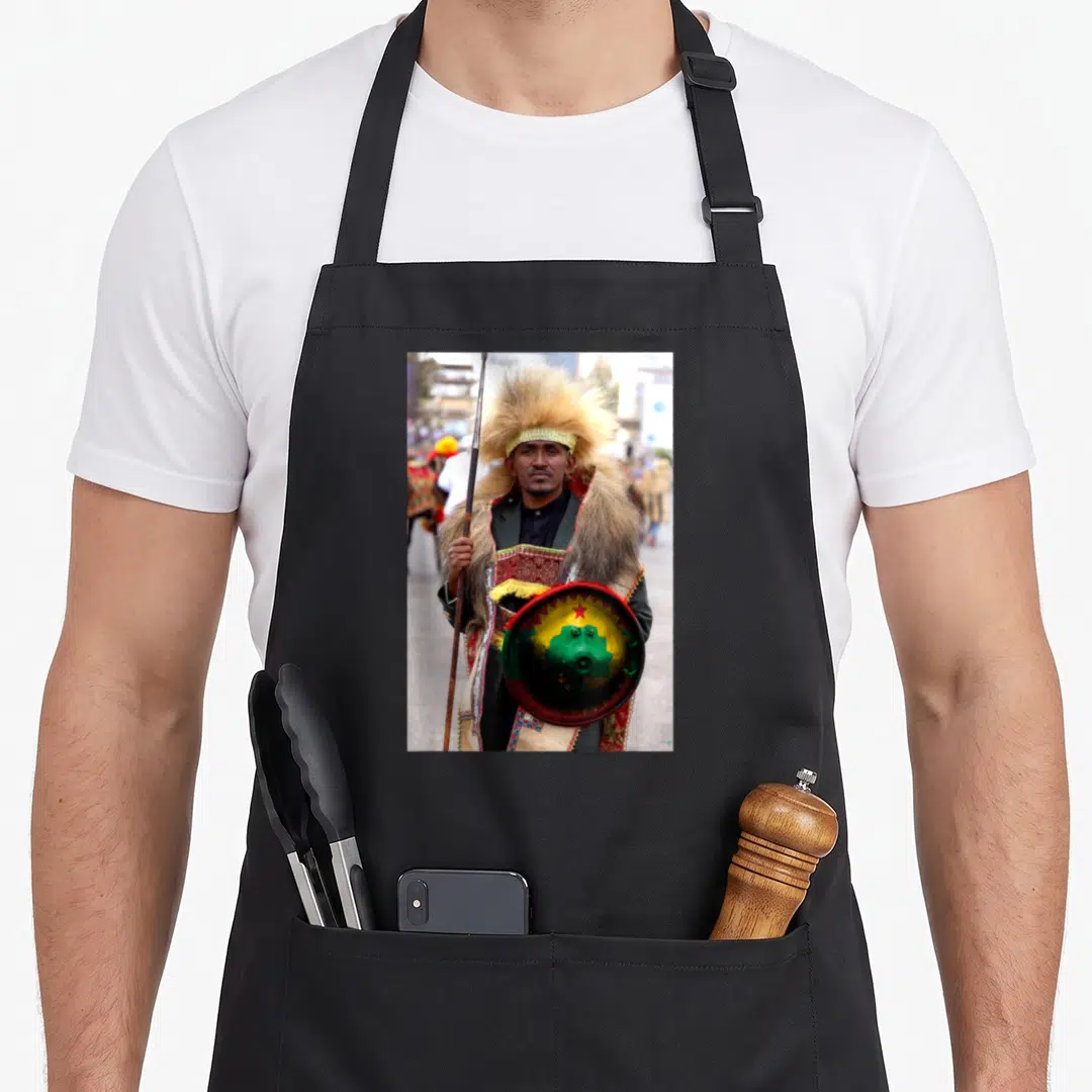 Oromo ethiopian boy apron - Man in traditional dress holding spear and shield with tan red green yellow colors — shown on a black cotton canvas apron against a white background