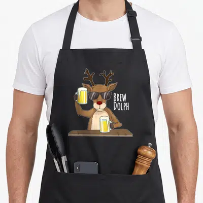 Brew holiday christmas aprons - Cartoon reindeer with sunglasses holding beer mugs next to stacked text — shown on a black cotton canvas apron against a white background