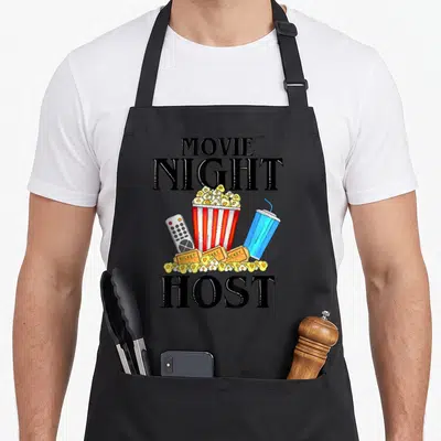 Host family aprons - Movie Night Host text surrounding a popcorn bucket, remote, blue drink, and tickets in cartoon — shown on a black cotton canvas apron against a white background