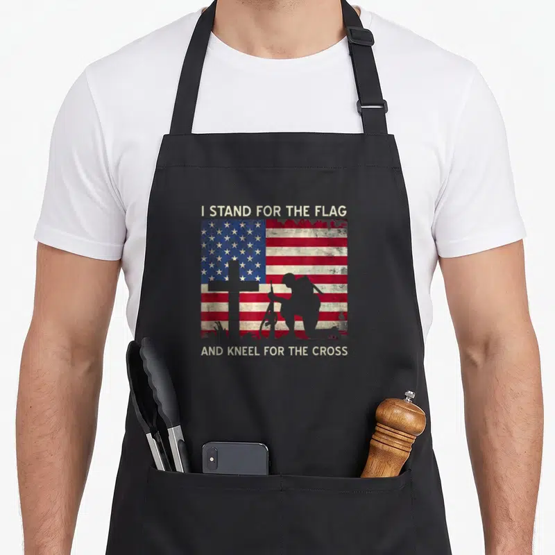 Flag kneel cross apron - Distressed USA flag with kneeling soldier silhouette and cross in grey, red, white, and blue — shown on a black cotton canvas apron against a white background