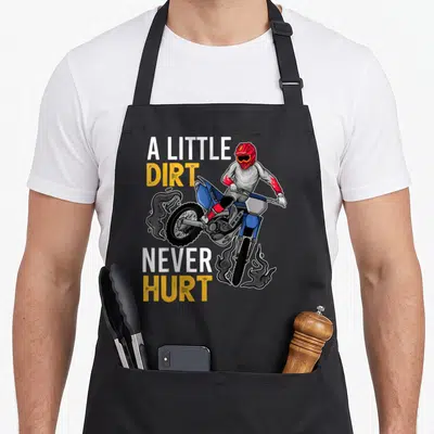 Motorbike bike apron - A LITTLE DIRT NEVER HURT text with cartoon motocross rider on blue bike — shown on a black cotton canvas apron against a white background