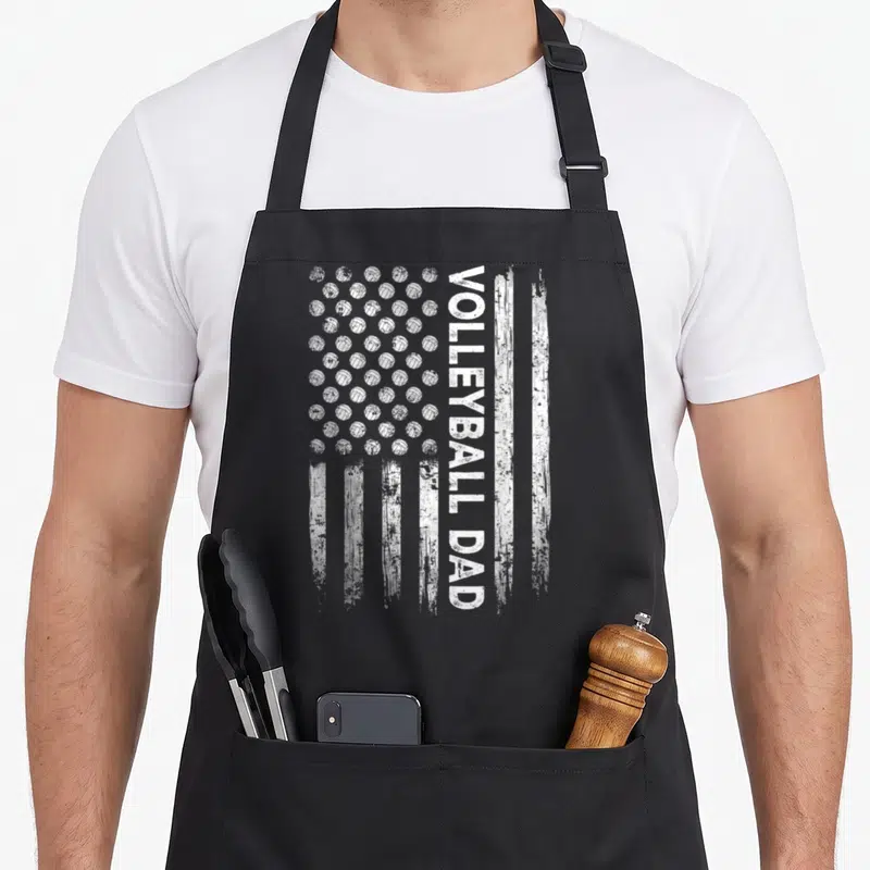 Volleyball father day apron - Distressed white USA flag with volleyball star grid and vertical VOLLEYBALL DAD text — shown on a black cotton canvas apron against a white background